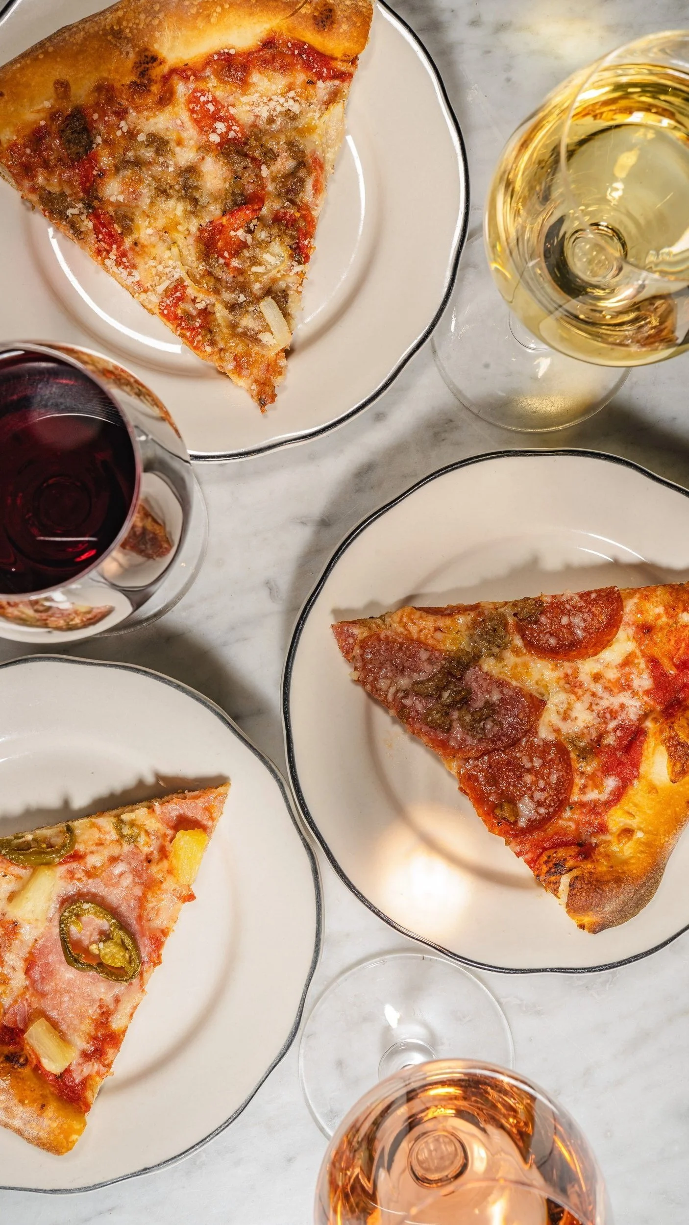 Slices of pepperoni pizza on white plates beside glasses of red and white wine on a marble table